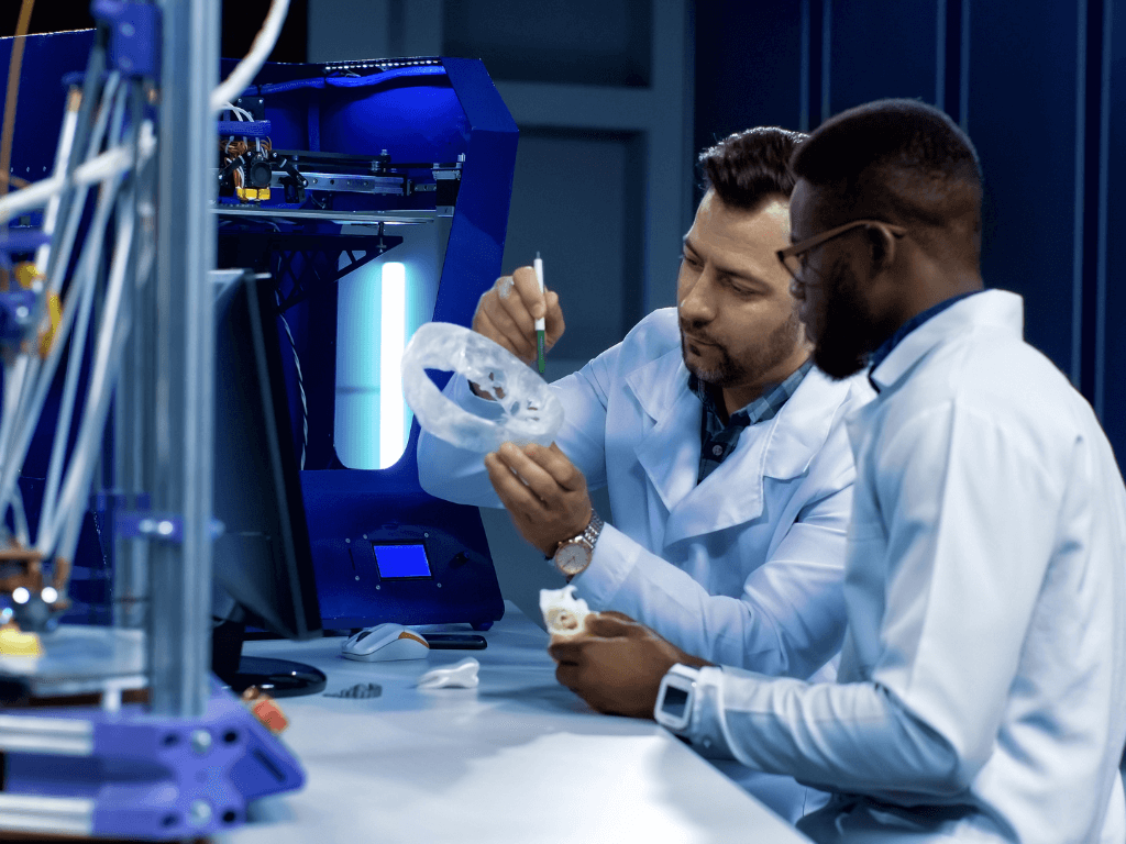 Two medical engineers in lab coats examine a 3D-printed prosthetic prototype at a desk with a 3D printer in the background.